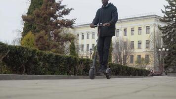 un joven afroamericano hombre en un negro suéter y negro pantalones y zapatillas paseos un eléctrico scooter abajo un callejón en el parque video