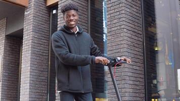 een jong Afrikaanse Amerikaans Mens in gewoontjes donker kleren met een scooter staat in de buurt een gebouw met een donker steen facade video