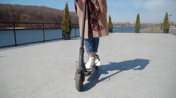 Young girl student in jeans and a coat rides on an electric scooter near the river. Close up view of the scooter and wheels from front video