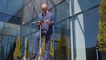 un afroamericano hombre en un clásico azul negocio traje soportes con un eléctrico scooter en el calle cerca un moderno espejo edificio y usos un teléfono inteligente fondo ver en lleno longitud video