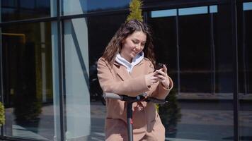 A young student girl in a sweater, jeans and a coat with an electric scooter stands near a stylish modern shopping center building and uses a smartphone. Front view video