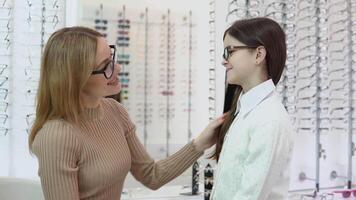 A blonde woman in a beige jacket and a dark-haired girl in a cute white cardigan and shirt pick up glasses for a child's vision correction at an eyewear store. Vision correction concept. Side view video