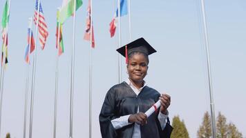 jovem africano americano fêmea graduado em pé dentro frente do a Câmera com uma diploma dentro dela mãos. a aluna carrinhos lado de fora com a internacional bandeiras em fundo. video