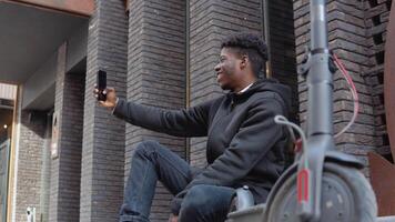 un joven africano americano hombre en un negro suéter pantalones y zapatillas se sienta en el acera cerca un edificio con un oscuro ladrillo fachada. el chico toma un selfie con un eléctrico scooter video