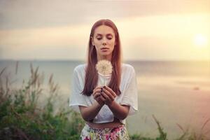 retrato de un morena joven mujer con un grande diente de león en un antecedentes de calentar puesta de sol. verano, al aire libre. foto