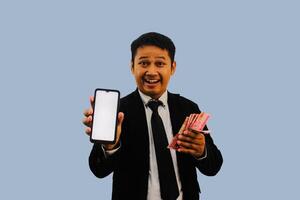 adulto asiático hombre sonriente contento mientras demostración blanco móvil teléfono pantalla y participación papel dinero foto