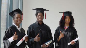 två ung afrikansk amerikan kvinnor och ett ung amerikan examen med diplom och böcker i deras händer är stående förbi de fönster. studenter är klädd i en festlig morgonrock och en mästare hatt video