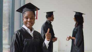 een jong Afro-Amerikaans vrouw staat in de voorgrond en looks Bij de camera met een diploma in haar handen. jong Afrikaanse Amerikaans Mens en vrouw communiceren met elk andere in de achtergrond. video