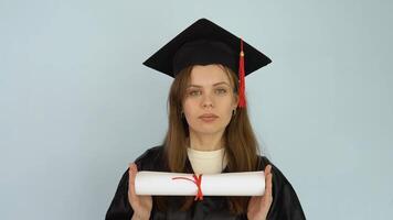 Young female student in a black gown and a master's hat holds in her hands in a horizontal position a diploma of higher education. White background video