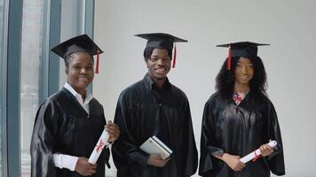 Tres afroamericano Universidad graduados con diplomas y libros en su manos estar por un ventana con un gris marco. estudiantes son vestido en un festivo túnica y un maestría sombrero video