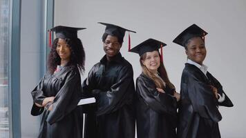 Two young women and one young African-American man and one young caucasian female university graduate with diplomas and books in their hands stand shoulder to shoulder by the window. video