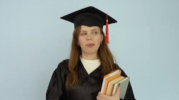 A young Caucasian woman in a black robe and a master's hat stands straight holding textbooks with one hand and looks at the camera. Portrait of half height on white background video