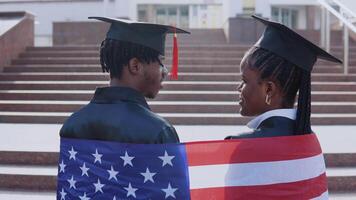 African American man and woman standing side by side with their backs to the camera. They have the British flag on their shoulders. A University building on background video