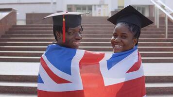 afrikansk amerikan man och kvinna stående sida förbi sida med deras ryggar till de kamera i svart klädnader och fyrkant hattar av examen studenter med de brittiskt flagga på deras axlar. ser till de kamera video