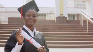 giovane africano americano femmina diplomato in piedi nel davanti di il telecamera con un' diploma e libri nel sua mani. il alunno è vestito nel un' nero accappatoio e un' piazza del maestro cappello e sta fuori. video