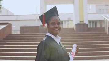 Young african american female graduate standing in front of the camera with a diploma and books in her hands. The student is dressed in a black robe and a square master's hat and stands outside. video