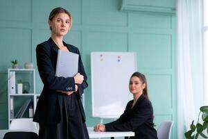 Two young women leaders dressed black suit in office one woman standing with document folder other woman sitting on the table looking at camera. Business meeting photo