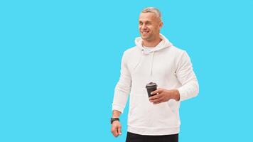 Men holding coffee in hand standing isolated blue background studio shoot. Caucasian handsome male dressed white hoodie looking away positive emotions teeth smiling. photo