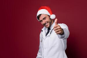 Young handsome doctor in white uniforme and Santa Claus hat standing in studio on red background smile and showing a thumbs up sign. photo