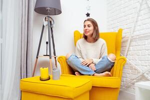 Enjoying leisure time at home. Beautiful young woman sitting in a soft yellow chair resting. Aromatic candles stand on the side photo