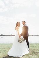 pareja vestida de boda con un ramo de flores y vegetación está en las manos contra el telón de fondo del campo al atardecer, la novia y el novio foto