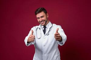 Young handsome modern doctor in a white medical gown stands in the studio on a red background. Student trainee of a medical university. photo