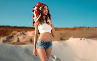 A beautiful young Caucasian girl in a white top and denim shorts on her head is wearing an Indian hat. Roach is in the desert. Happy holiday mood. photo