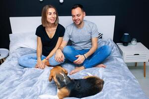 Young couple sitting on a bed in the bedroom, hugging. Near them is their dog photo