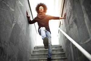 hermoso joven hombre de mezclado carrera con un Corte de pelo en de moda ropa, pantalones y un bufanda posando en contra el antecedentes de un pared y escalera hecho de cerámico granito losas foto