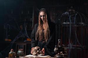 Halloween concept. Witch portrait close up with dreadlocks dressed black hood standing dark room with cage on background photo