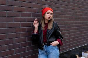 Beautiful young stylishly dressed Caucasian girl smokes cigarette on the street. photo