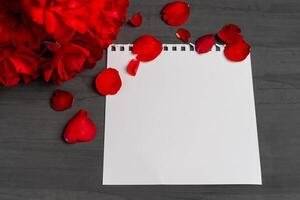 Empty sheet of paper for text next to a bouquet and red rose petals against a dark background. photo