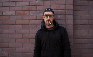 Handsome young unshaven man black hoodie and bandana stands backyard near brick walls summer day. photo