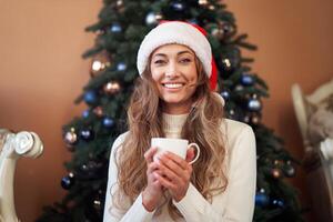 Christmas. Woman dressed white sweater Santa hat and jeans sitting on the floor near christmas tree with present box photo