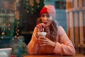 Beautiful caucasian blonde woman in red knitted hat sitting behind cafeteria window drinking a warm latte on a wonderful Christmas day. photo