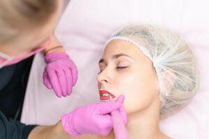 Cosmetology cabinet client lies on couch. Beautician applies marking on lips. photo