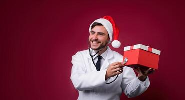 Young handsome doctor in white uniforme and Santa Claus hat standing in studio on red background smile and finger in camera photo