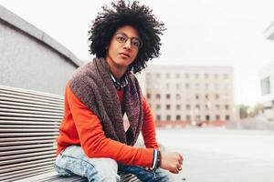 A handsome young Egyptian curly guy sitting on a bench dressed in a bright orange sweater and jeans. The student walks in the break between the lessons. Cute hipster guy photo