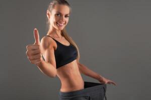 Slim young woman wearing old big pants, showing successful result of a diet - weight loss, slimming concept. gray background photo