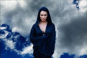 Young beautiful woman with black hair and in the dark blue cloak with hood at the sky background photo