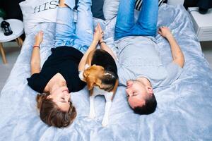 Young couple sitting on a bed in the bedroom, hugging. Near them is their dog photo