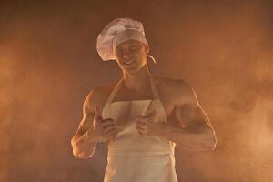 retrato de muscular cocinero vistiendo blanco delantal y cocinero sombrero, demostración pulgar arriba en ahumado antecedentes foto