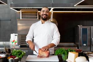 Handsome young African chef standing in professional kitchen in restaurant preparing a meal of meat and cheese vegetables. photo