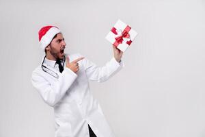 Young handsome doctor in white uniforme and Santa Claus hat standing in studio on white background smile and Shows a finger at the gift box. photo