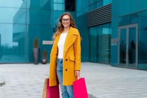 Business woman with shopping bags dressed yellow coat walking outdoors corporative building background photo