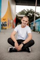 Young Man Smiling, Sitting on Ground, Hands on Knees - Positive Expression, Casual Outdoor Portrait - Happy Young Male Looking at Camera, Relaxed Pose - Lifestyle Concept photo