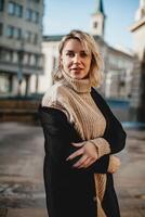 sonriente mujer en negro Saco y marrón suéter poses en urbano acera. confidente hembra modelo sonrisas para cámara en ciudad calle escena. elegante Moda retrato de mujer en elegante ropa de calle en acera foto