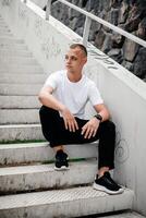 Young Man in Casual Attire Sitting on Stairs. Looking at Camera. Relaxed Mood Portrait of Stylish Guy in White Shirt and Black Pants, Wearing Watch. Urban Lifestyle Concept, Casual Fashion Photography photo