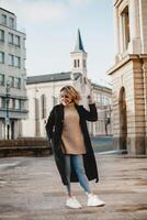 Stylish Woman in Black Coat and Jeans Smiling on Sidewalk - Urban Lifestyle, Happy Female Enjoying Cityscape - Casual Fashion and Joyful Mood in Urban Environment, Building Background, City Life. photo