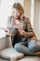 relajado mujer leyendo revista en amortiguar - casual ocio hora con impreso medios de comunicación - joven dama disfrutando tranquilo momento en elegante chaqueta y vaqueros, acogedor estilo de vida concepto. foto